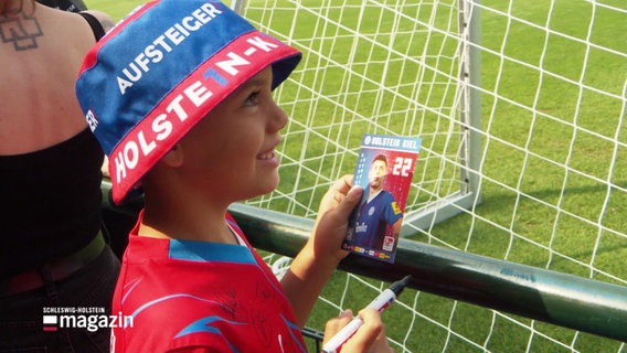 Ein Kind mit Holstein-Kiel Fankleidung und Autogrammkarte schaut den Spielern beim Training zu. © Screenshot 