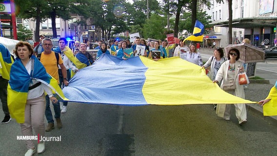 Menschen auf einer Demo tragen eine große Flagge der Ukraine zwischen sich. © Screenshot 