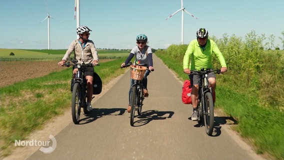 Zwei Radfharer und eine Radfahrerin fahren über einen Feldweg. Im Hintergrund: Blauer Himmel, Grüne Felder und Windräder. © Screenshot 