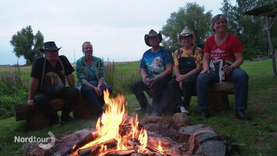 Eine Gruppe Frauen und Männer, einige tragen Cowboyhüte, sitzt um ein Lagerfeuer. © Screenshot 