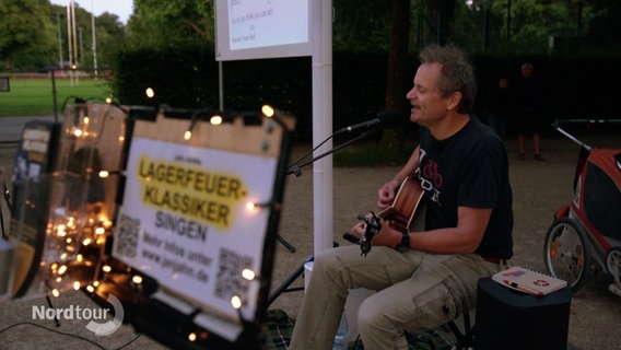 Jan Jahn sitzt singend mit seiner Gitarre am Wegesrand. Vor ihm steht ein Schild mit der Aufschrift "Lagerfeuer-Klassiker". © Screenshot 