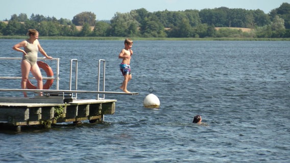 Ein kleiner Junge hüpft auf dem Sprungbrett einer schwimmenden Plattform auf einem Badesee auf und ab. © Screenshot 