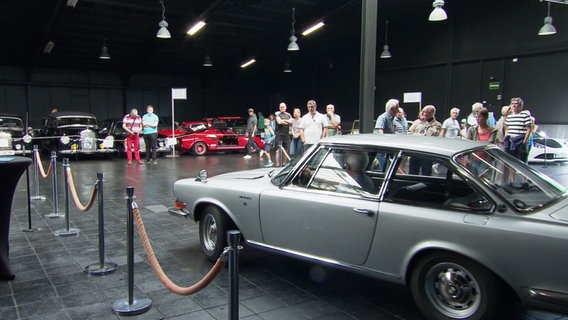 Ein Oldtimer wird in eine Halle gefahren, in der bereits interessierte Autofans warten. © Screenshot 