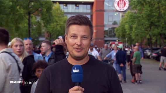 NDR Reporter Sebastian Rieck steht mit einem Mikrofon vor dem Millerntor-Stadion in Hamburg © Screenshot 