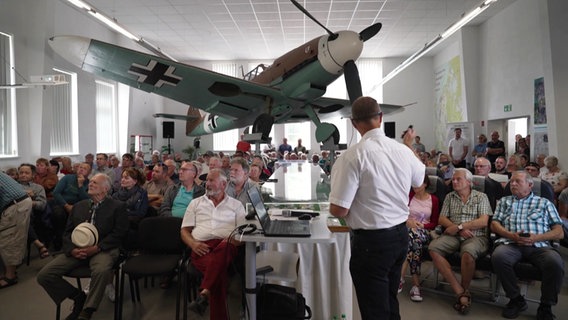 Eine Versammlung in einer Halle, in der ein historisches Flugzeug hängt. © Screenshot 