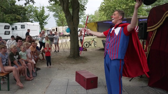 In Clownskostüm und mit rotem Umhang lüpft Holger Pautz theatralisch seinen Hut vor einerm Kinderpublikum. © Screenshot 