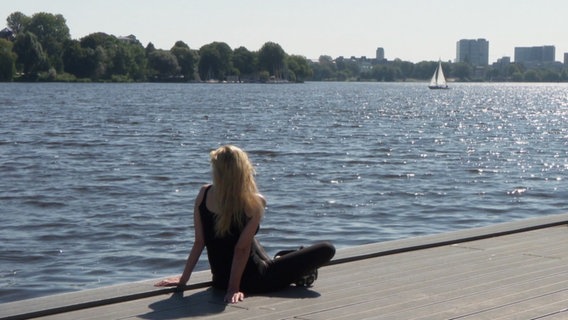 Eine Person sitzt am Wasser, im Hintergrund ein kleines Segelschiff. © Screenshot 