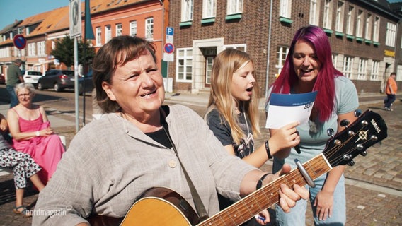 Danie Behm, eine Frau mit pink gefärbten Haaren, singt mit einer Frau, die Gitarre spielt und einem Mädchen. © Screenshot 