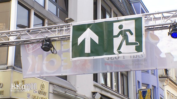 An einem Querbalken hängt auf einem Straßenfest ein Fluchtwege-Schild. © Screenshot 