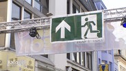 An einem Querbalken hängt auf einem Straßenfest ein Fluchtwege-Schild. © Screenshot 