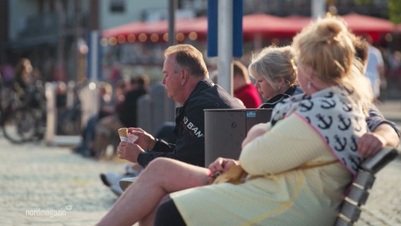 Auf einer Bank an einer Promenade sitzen mehrere Menschen. Ein Mann unter ihnen isst ein Eis aus der Waffel. © Screenshot 