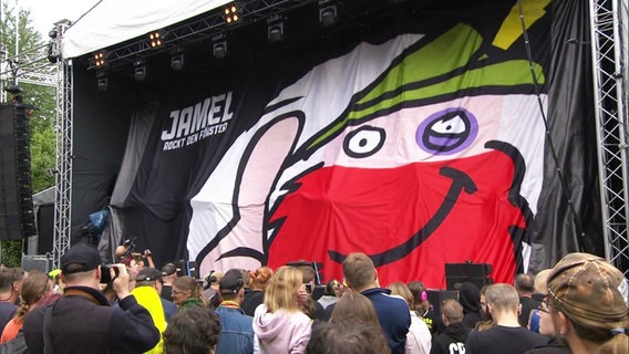 Festivalpublikum steht vor einer Bühne, die mit einem Vorhang verhangen ist, auf dem das "Jamel Rockt den Förster" Logo prangt. © Screenshot 