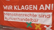 Auf einem von Demonstrierenden gehaltenen Transparent steht "Wir klagen an! Menschenrechte sind #unverhandelbar". © Screenshot 