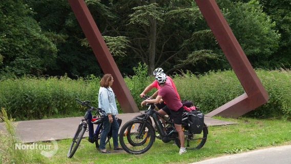 Eine Frau und zwei Männer stehen mit ihren Fahrrädern vor einer Skulptur aus Stahl. © Screenshot 