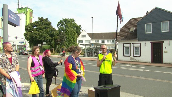 Die Organisatoren vom ersten CSD in Burgdorf bei einer Rede während eines Demozuges. Ein Mann in gelber Warnweste hält eine abgebrannte Regenbogenflagge empor. Neben ihm steht eine Frau, die eine intakte Flagge über ihren Schultern trägt. © Screenshot 