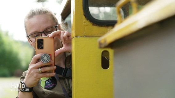 Eine Frau lehnt sich aus der Tür einer gelben Förderbahn und richtet ihr Smartphone zum Fotografieren auf die Kamera des Betrachtenden. © Screenshot 