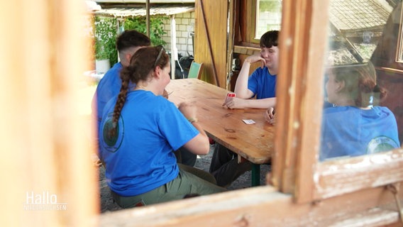 Blick durch ein offenes Fenster mit Holzrahmen. Jugendliche sitzen um einen Holztisch und spielen Karten. Sie alle tragen ein blaues T-Shirt. © Screenshot 