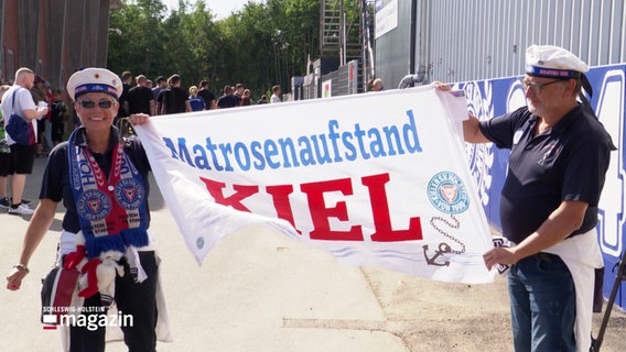 Zwei Holstein-Kiel-Fans halten ein Banner hoch. Darauf steht "Matrosenaufstand Kiel". Sie beide tragen einen Matrosenhut auf dem Kopf, die Frau links ist noch mit Holstein-Kiel-Fans geschmückt. © Screenshot 