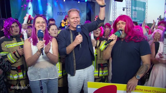 Von links nach rechts: Dania Behm, eine Frau mit pinken, lila Haaren, Thilo Tautz, und Stefan Kuna mit einer pinkfarbenen Langhaarperücke, jubeln in die Kamera. © Screenshot 