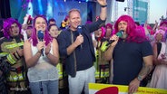 Von links nach rechts: Dania Behm, eine Frau mit pinken, lila Haaren, Thilo Tautz, und Stefan Kuna mit einer pinkfarbenen Langhaarperücke, jubeln in die Kamera. © Screenshot 