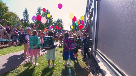 Kinder stehen mit dem Rücken zur Kamera. Auf dem Rücken tragen sie bunte Schulranzen. In die Luft steigen Luftballons in verschiedenen Farben. © Screenshot 