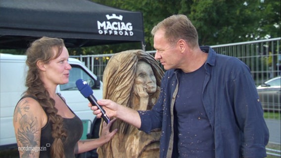 Thilo Tautz interviewt Kettensägen-Künstlerin Sabrina Bartz. Hinter beiden steht eine Statue aus Holz, die Rapunzel darstellen soll. © Screenshot 