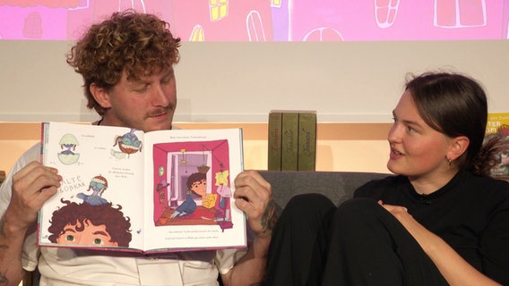 Malte Zierden hält ein aufgeschlagenes Kinderbuch in den Händen. © Screenshot 