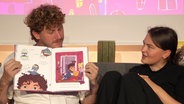 Malte Zierden hält ein aufgeschlagenes Kinderbuch in den Händen. © Screenshot 