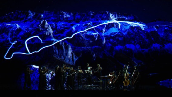 Ein klassisches Konzert vor beieindruckender Videokulisse, es sind Berge und ein blaues Licht zu sehen. © Screenshot 