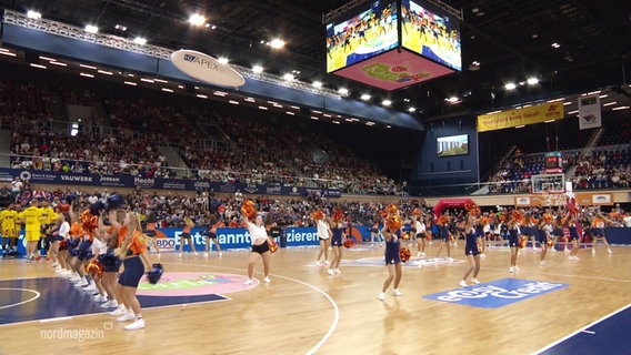 In einer Basketball-Arena führt ein Cheerleading-Team eine Choreographie auf. © Screenshot 