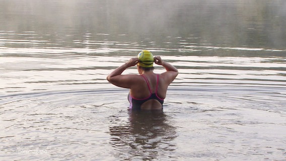 Eine Frau im Badeanzug und mit Badekappe steht mit dem Rücken zur Kamera bis zum Bauch im Wasser eines Sees. © Screenshot 