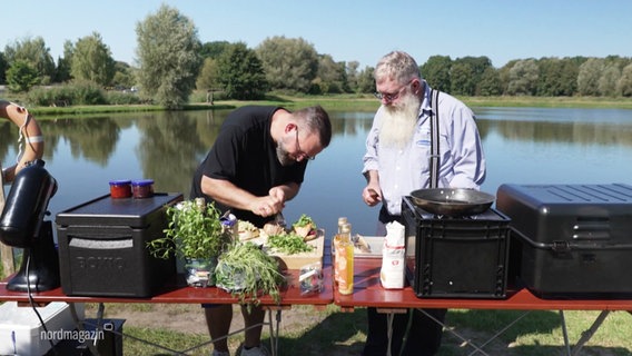 Zwei Personen kochen an einem Tisch vor einem See. © Screenshot 