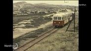 Ein sogenannter "Schienenbus", ein Kleinzug bestehend aus zwei Waggons, fährt auf einer historischen Filmaufnahme durch eine Dünenlandschaft. © Screenshot 