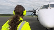 Ein Mitglied des Bodenpersonals an einem Flughafen steht in gelb-leuchtender Arbeitskleidung und Hörschutz vor einer kleineren Passagiermaschine. © Screenshot 