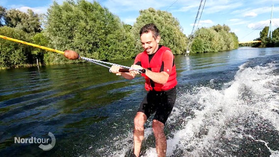 Ein Mann in Badeshorts und mit roter Schwimmweste fährt auf einem See Wasserski in einer Anlage. © Screenshot 