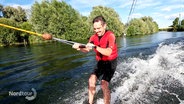 Ein Mann in Badeshorts und mit roter Schwimmweste fährt auf einem See Wasserski in einer Anlage. © Screenshot 