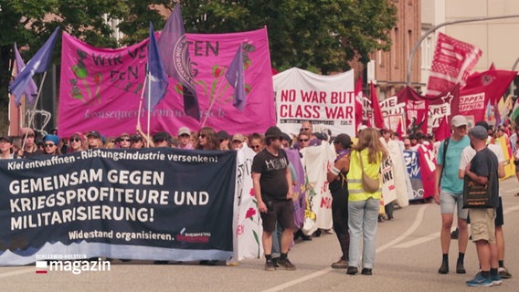 Menschen demonstrieren. Sie halten Banner und Fahnen empor, auf einigen stehen ablehnende Parolen gegen die Krigsindustrie. © Screenshot 