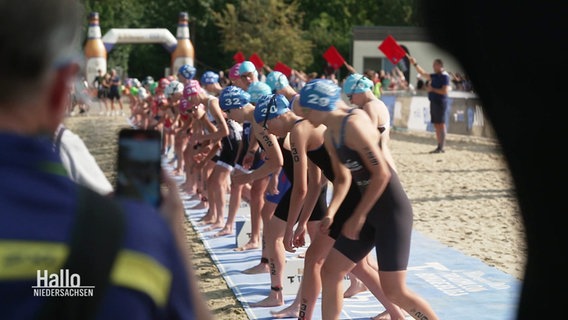 An einer Startlinie an einem Strand stehen viele Triathleten vor dem Startschuss. © Screenshot 