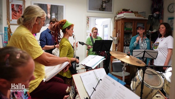 Eine Gruppe von Menschen mit bunten Klamotten macht als Band zusammen in einem Raum Musik. © Screenshot 