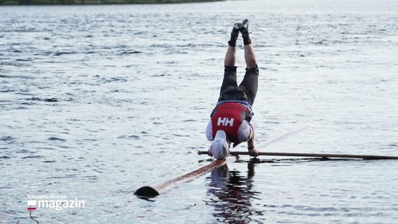 Ein Mann macht einen Unterarmstand auf einem Baumsamm, der im Wasser treibt. © Screenshot 