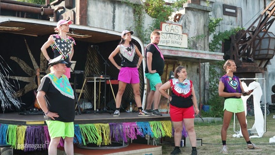 Auf einer kleineren Bühne auf einem Theaterfestival stehen Darstellende in neon-bunten Outfits. © Screenshot 