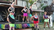 Auf einer kleineren Bühne auf einem Theaterfestival stehen Darstellende in neon-bunten Outfits. © Screenshot 