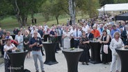 Bei einer Preisverleihung stehen viele Zuschauende an Stehtischen in einem Parkgelände. © Screenshot 