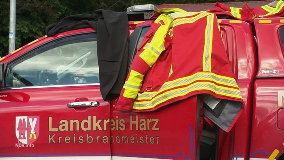 Eine Arbeitsjacke liegt auf einem roten Auto mit der Aufrschrift: Landkreis Harz, Kreisbrandmeister. © Screenshot 