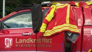 Eine Arbeitsjacke liegt auf einem roten Auto mit der Aufrschrift: Landkreis Harz, Kreisbrandmeister. © Screenshot 