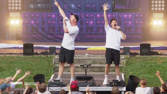 Mitglieder der Band "Deine Freunde" am Bühnenrand beim Konzert im Stadtpark: Sie singen ins Mikro und recken die Arme in die Luft. © Screenshot 