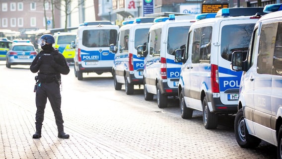 Ein einzelner Polizist steht breitbeinig auf einer Straße, an deren Rand zahlreiche Mannschaftswagen parken. © Screenshot 