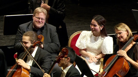 Ein Mann und eine Frau sitzen als Zuschauer mitten im Orchester bei den Cellisten. © Screenshot 