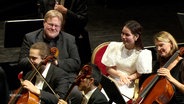 Ein Mann und eine Frau sitzen als Zuschauer mitten im Orchester bei den Cellisten. © Screenshot 