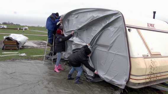 Drei Personen versuchen bei starkem Wind das Vorzelt eines Campinganhängers aufzubauen. © Screenshot 